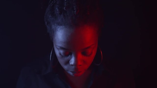 Tilt down low-key portrait shot with red and blue lighting: confident black woman with braids in hair bun and hoops earrings looking up and staring at camera with daring expression