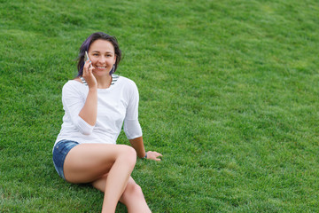 Happy beautiful young woman sitting on grass and using smartphone