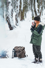 Fototapeta na wymiar Girl standing near camp fire with a cup of hot coffee in the snowy forest. Concept adventure active vacations outdoor. Winter camping. vertical photo
