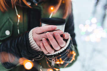 Fototapeta na wymiar Close up view. Girl hands with a cup of hot coffee, tea, in the snowy forest. Concept adventure active vacations outdoor. Winter camping, copy space, colorful christmas lights