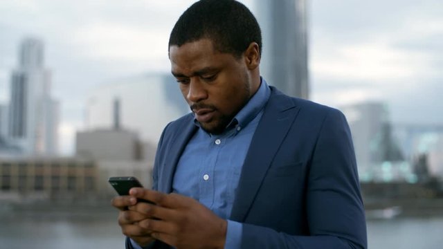 Panning of African businessman in blue jacket standing near river and office buildings, holding cellphone and chatting with someone