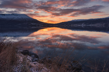 Fototapeta na wymiar Beautiful orange sunset sunset landscape with pink clouds in a fjord reflection in Norway near the city of Tromso with hoarfrost on the rocks near the ocean