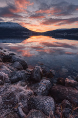 Beautiful orange sunset sunset landscape with pink clouds in a fjord reflection in Norway near the city of Tromso with hoarfrost on the rocks near the ocean