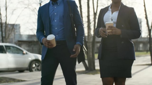 Tilt up of African businessman walking with African businesswoman, holding coffee paper cup and talking