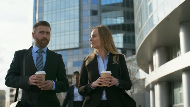 Medium shot of Caucasian businessman and Caucasian business lady walking, holding paper coffee cups and having conversation