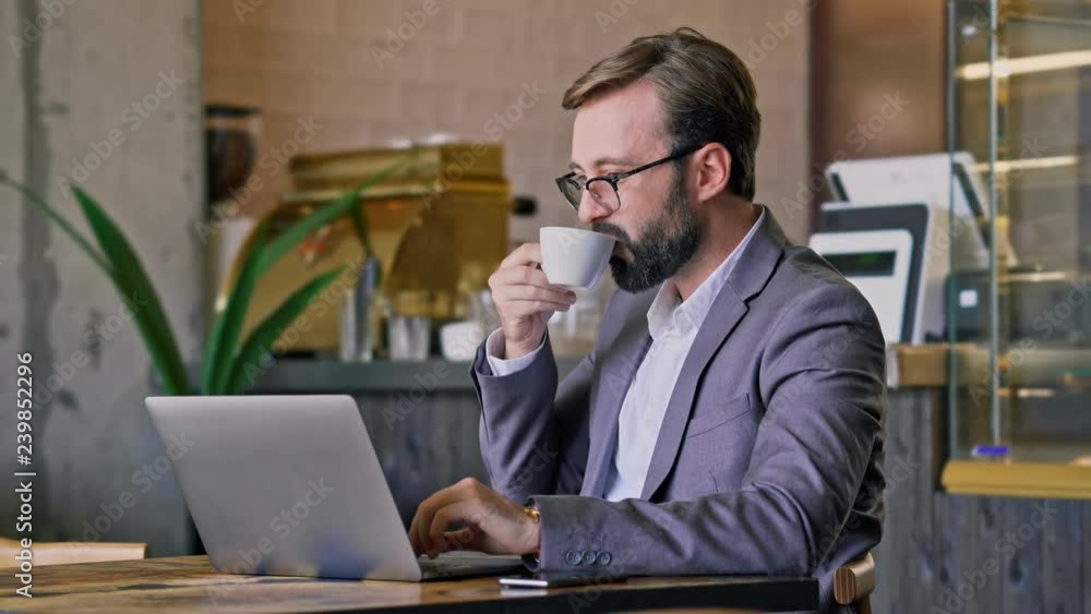 Wall mural Cheerful bearded business man in eyeglasses using laptop computer and drinking coffee while sitting by the table at cafe