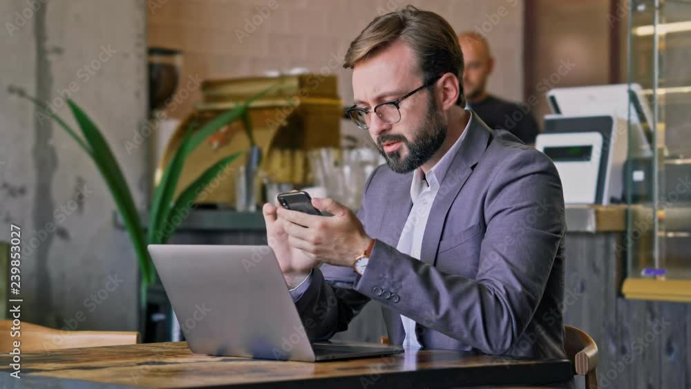 Wall mural Serious bearded business man in eyeglasses using smartphone while sitting by the table at cafe
