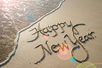 Happy New Year message handwritten in smooth sand with sunrise lens flare over oncoming wave on the beach