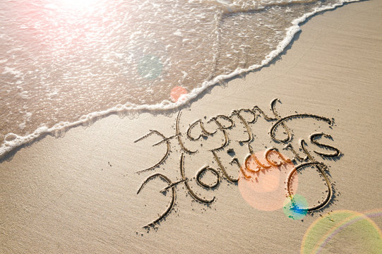 Happy Holidays message handwritten in smooth sand with an oncoming wave in the lens flare of the tropical sun
