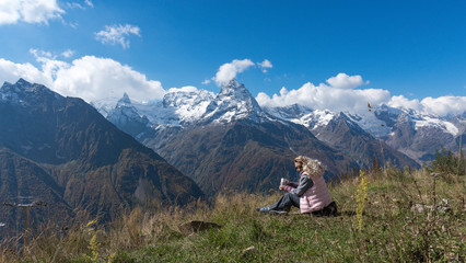 Fototapeta premium woman traveler drinks coffee with a view of the mountain landscape. A young tourist woman drinks a hot drink from a cup and enjoys the scenery in the mountains. Trekking concept.