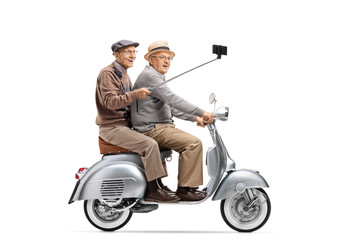 Two senior men riding on a vintage scooter taking a selfie with a stick