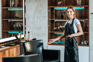beautiful young hairdresser showing empty chair and smiling at camera in beauty salon