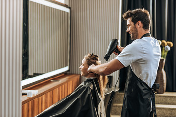 side view of smiling young hairstylist drying hair to happy girl in beauty salon