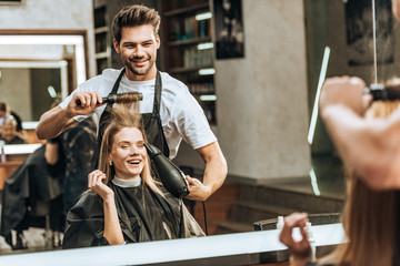 Smiling young hairstylist combing and drying hair to happy young woman in beauty salon