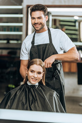 smiling hairstylist using hair straightener and happy girl looking at mirror in beauty salon