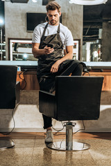 handsome young hairstylist in apron using smartphone in beauty salon