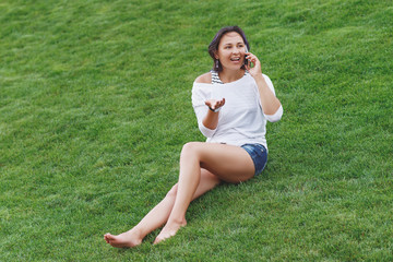 Happy beautiful young woman sitting on grass and using smartphone