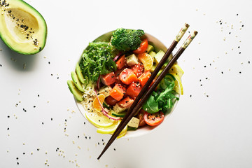 poke bowl with salmon, avocado, cucumber, arugula, broccoli, rice, carrot and sweet onions with chuka salad, with chopsticks, avocado isolated over white background. top view