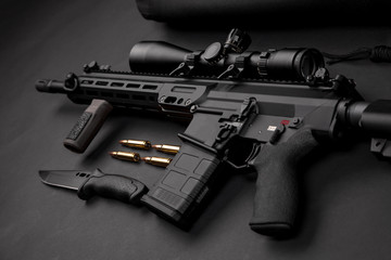 modern automatic rifle with an optical sight lies on a dark background and next to the knife and...