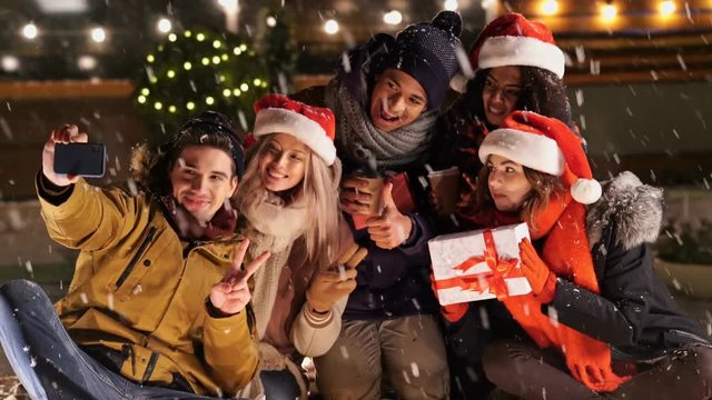 Multiethnic happy friends in warm clothes making selfie on smartphone in snow street
