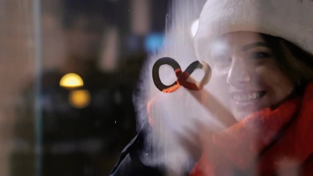 Happy caucasian lady in warm clothes drawing infinity sign on misted glass and showing heart gesture in snow street