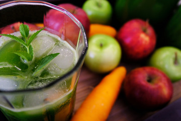 A glass of ice cold freshly squeezed fruit and vegetable juice, with some fruits and veggies in the background. A mint sprig is sitting on top of the drink.