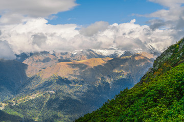 Naklejka premium Mountain landscape. Warm autumn Sunny day. The slopes of the mountains covered with autumn woods. Clouds above mountain peaks. The snow on the tops