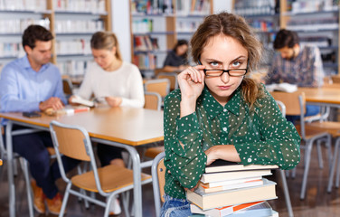 Annoyed girl sitting with pile of books in high school library on background of working fellow students