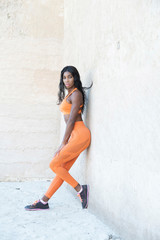 African American Black athletic model wearing bright orange sports leans against a concrete wall with with her foot on the wall 