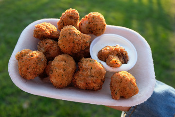 Deep fried mushrooms in batter with parsley and seasoning being held in a generic take out container. Mayo sauce with dill herb dip is on the side, slightly dripping. Angled view close up.