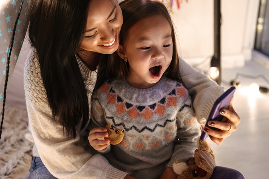 Happy Young Asian Woman With Her Little Daughter Girl Having Fun On Floor Using Mobile Phone. Christmas Concept.