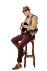 Fototapeta premium side view of mixed race male musician in hat and eyeglasses playing on acoustic guitar while sitting on chair isolated on white