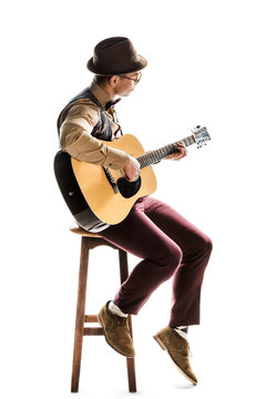 young mixed race male musician in hat and eyeglasses playing on acoustic guitar while sitting on chair isolated on white