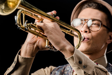 close up view of mixed race male jazzman in hat and eyeglasses playing on trumpet isolated on black