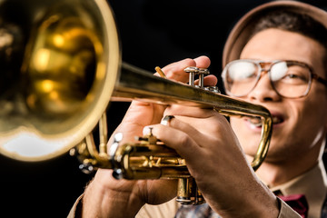 selective focus of male jazzman in hat and eyeglasses playing on trumpet isolated on black