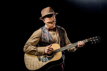 focused mixed race male musician in eyeglasses and hat playing on acoustic guitar isolated on black