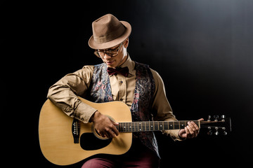 young handsome man in eyeglasses and hat playing on acoustic guitar isolated on black