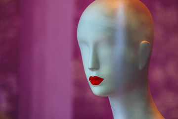 A bold white female mannequin with no eyes and red lips in a showcase of the fashion store on a black background.