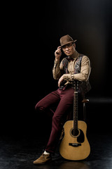 young man in stylish hat posing with acoustic guitar while sitting on chair on black