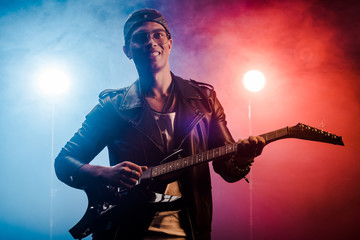 Obraz premium laughing young male musician in leather jacket performing on electric guitar on stage with smoke and dramatic lighting