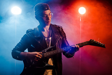 Obraz premium focused male musician in leather jacket performing on electric guitar on stage with smoke and dramatic lighting