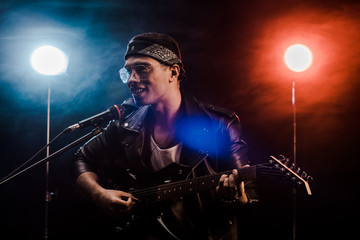 smiling male musician singing in microphone and playing on electric guitar on stage during rock concert