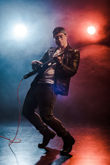 handsome stylish rocker in leather jacket performing on electric guitar on stage with smoke and dramatic lighting