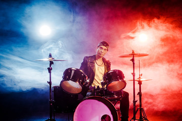 confident mixed race male musician sitting behind drum set on stage with spotlights