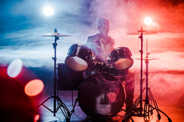male musician in leather jacket playing drums during rock concert on stage with smoke and spotlights