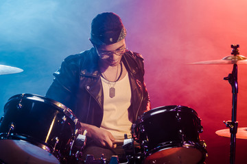 Fototapeta na wymiar smiling young male musician in leather jacket playing drums during rock concert on stage with smoke and dramatic lighting