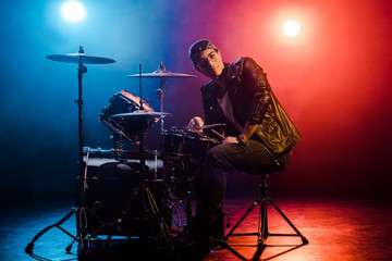 Fototapeta na wymiar young mixed race male musician sitting behind drum set on stage with spotlights and smoke