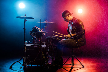 side view of male musician in leather jacket playing drums during rock concert on stage with smoke...