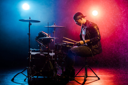 Side View Of Male Musician In Leather Jacket Playing Drums During Rock Concert On Stage With Smoke And Spotlights