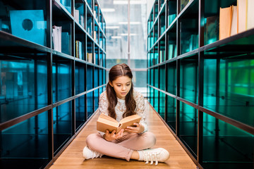 adorable concentrated schoolgirl reading book on floor in library